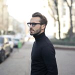 Home A fashionable man in a black top and glasses poses in an urban street with parked cars.